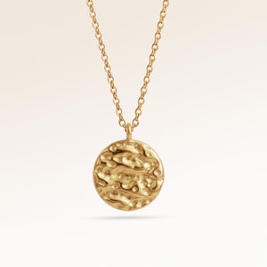Close up af minimalistisk rund vedhæng i guld med blødt smeltet udtryk og plads til indgravering bagpå