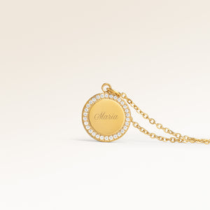 Close up af minimalistisk rund vedhæng i guld med hvide zirkoner langs kanten og plads til indgravering på forsiden